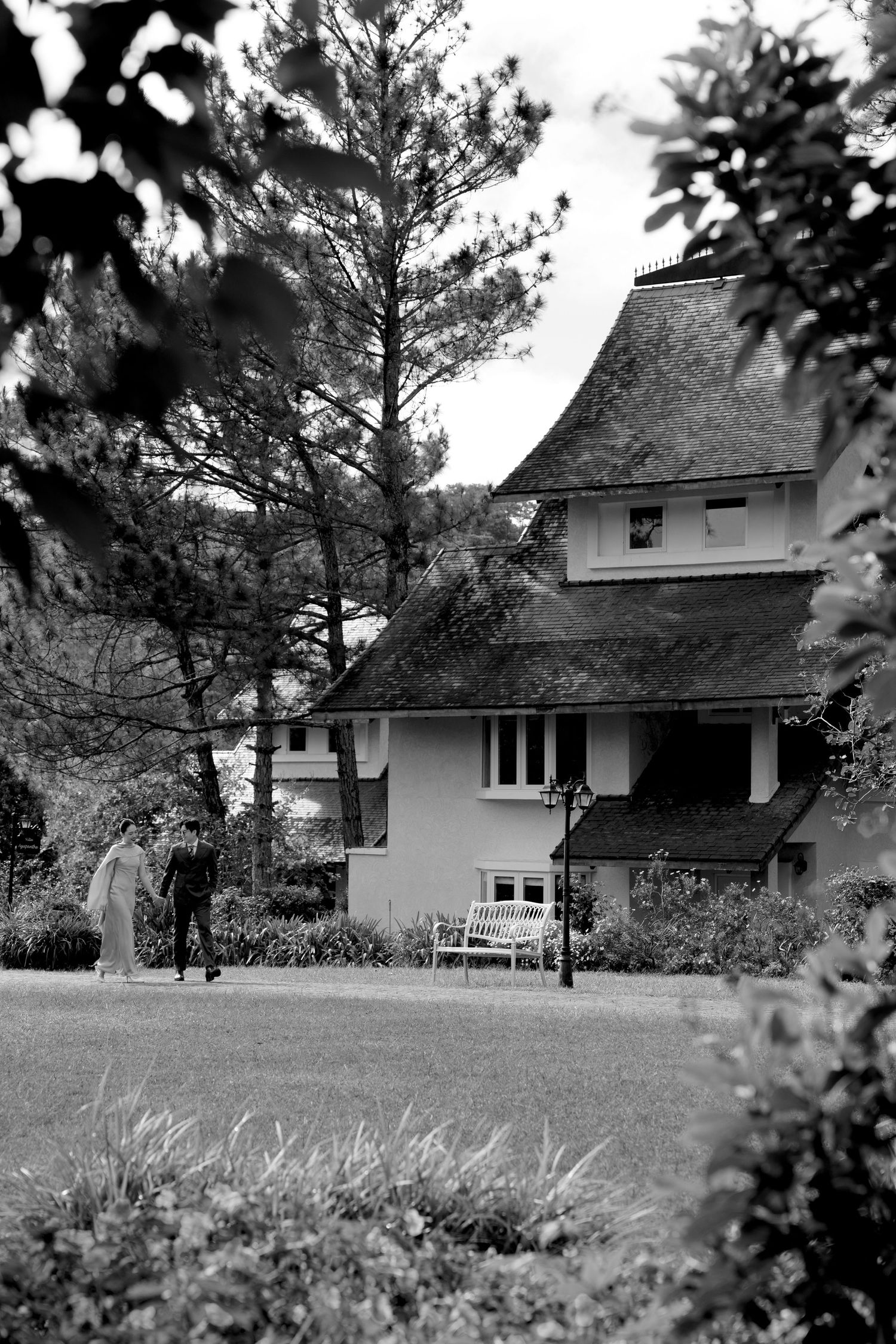 Black and white photo of a couple near a charming house surrounded by tall pine trees and lush garden.