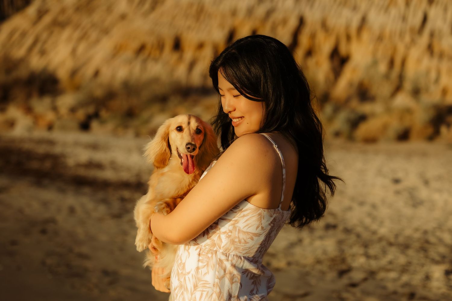 A smiling woman holds an adorable golden dachshund puppy on a sunlit beach at golden hour.