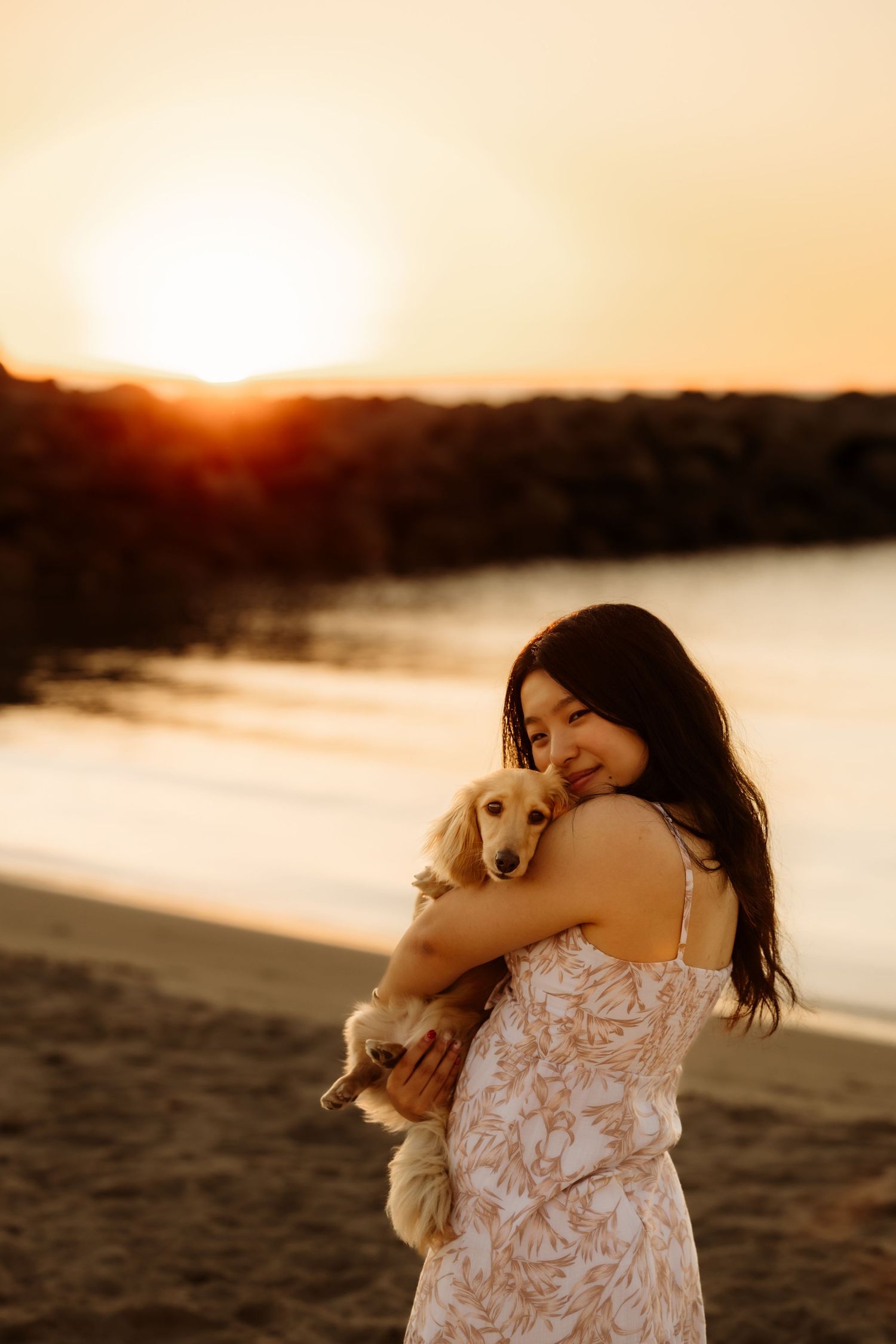 Woman in floral dress holding a golden dachshund on a sandy beach at golden sunset.
