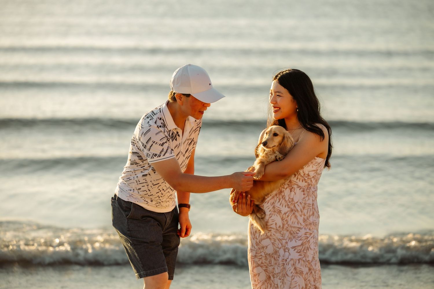 Couple smiling and holding a small dog together on a sunny beach with ocean waves in the background.
