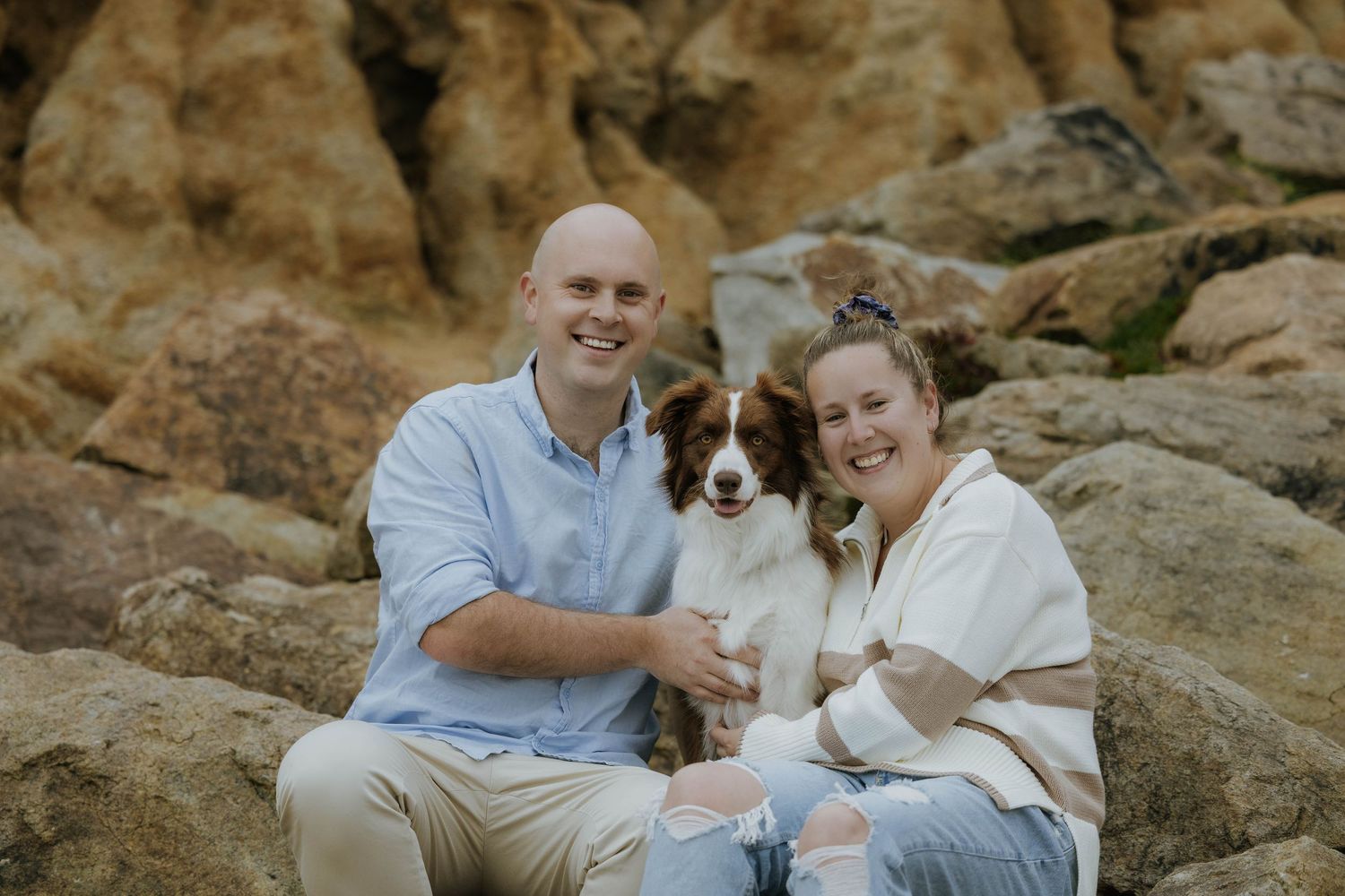 A couple and their dog sit together on rocky coastal cliffs during a romantic photoshoot at sunset.