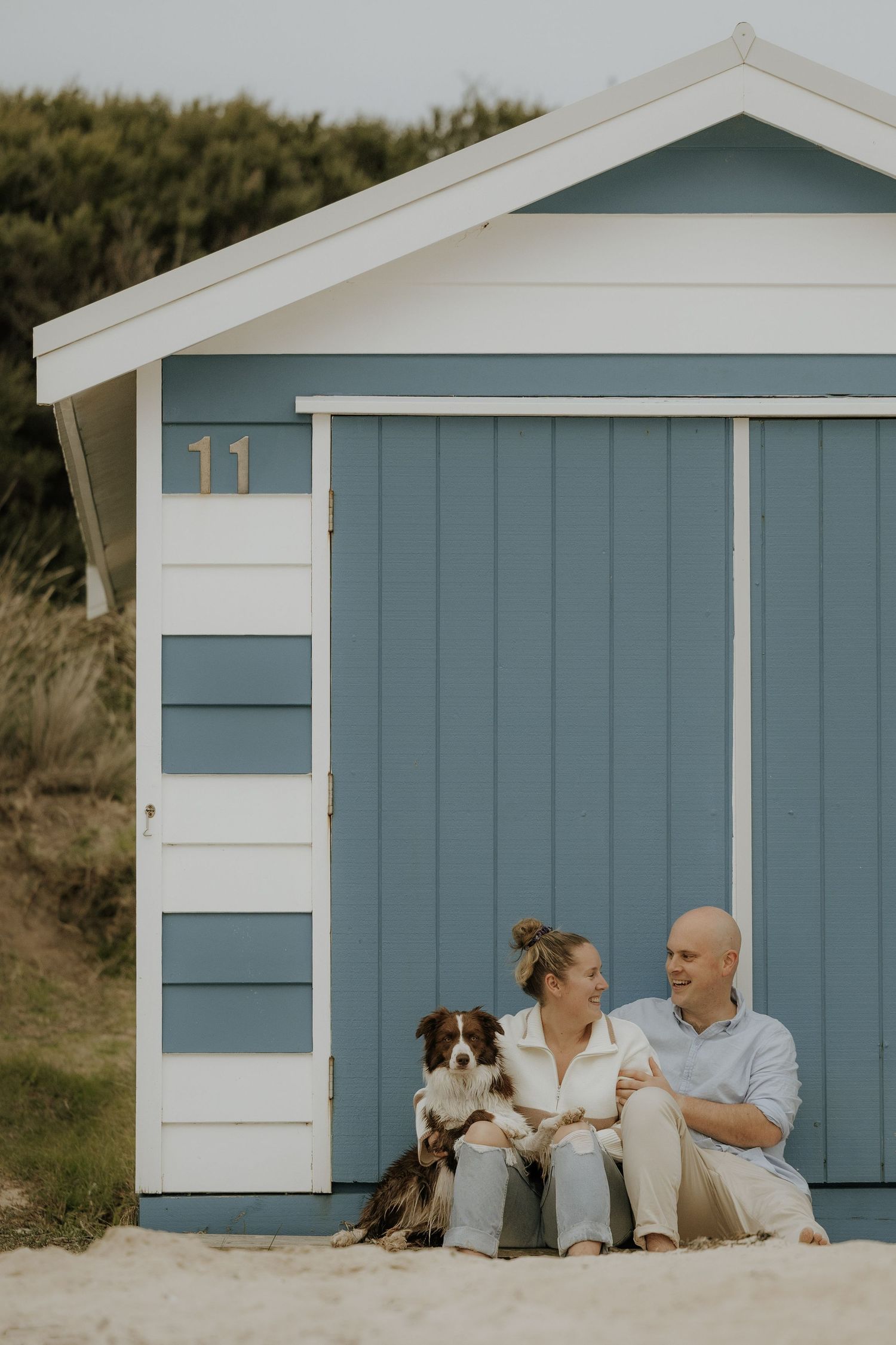 A couple and their dog relax in front of blue and white beach huts on a sunny day.