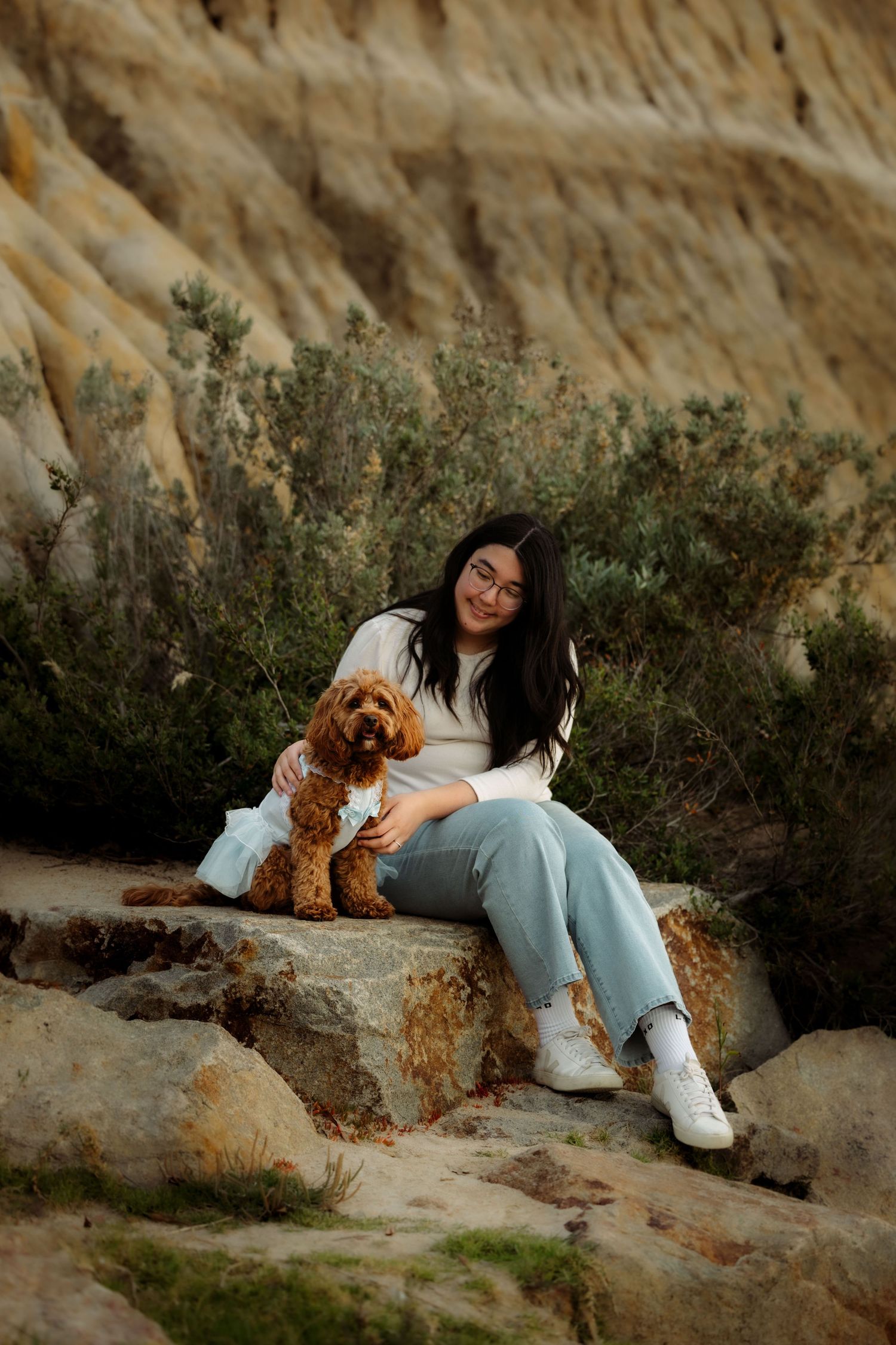 Young woman with dark hair sits on rocks with her fluffy golden Cavapoo dog in a scenic canyon landscape.