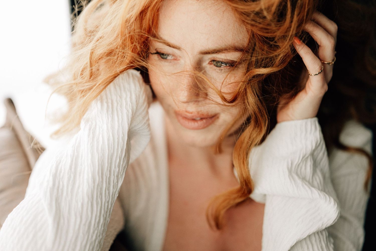 Person with wavy blonde hair wearing a white sweater smiles warmly while sitting indoors in natural light.