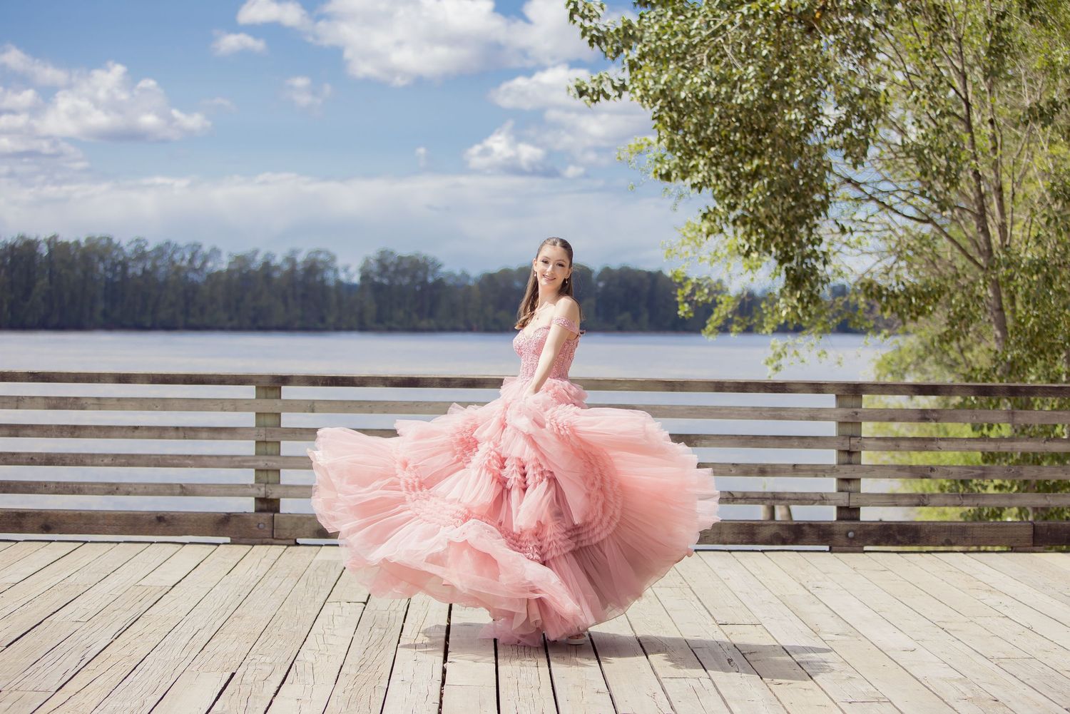 A flowing pink tulle gown twirls gracefully on a wooden deck overlooking a serene natural landscape.