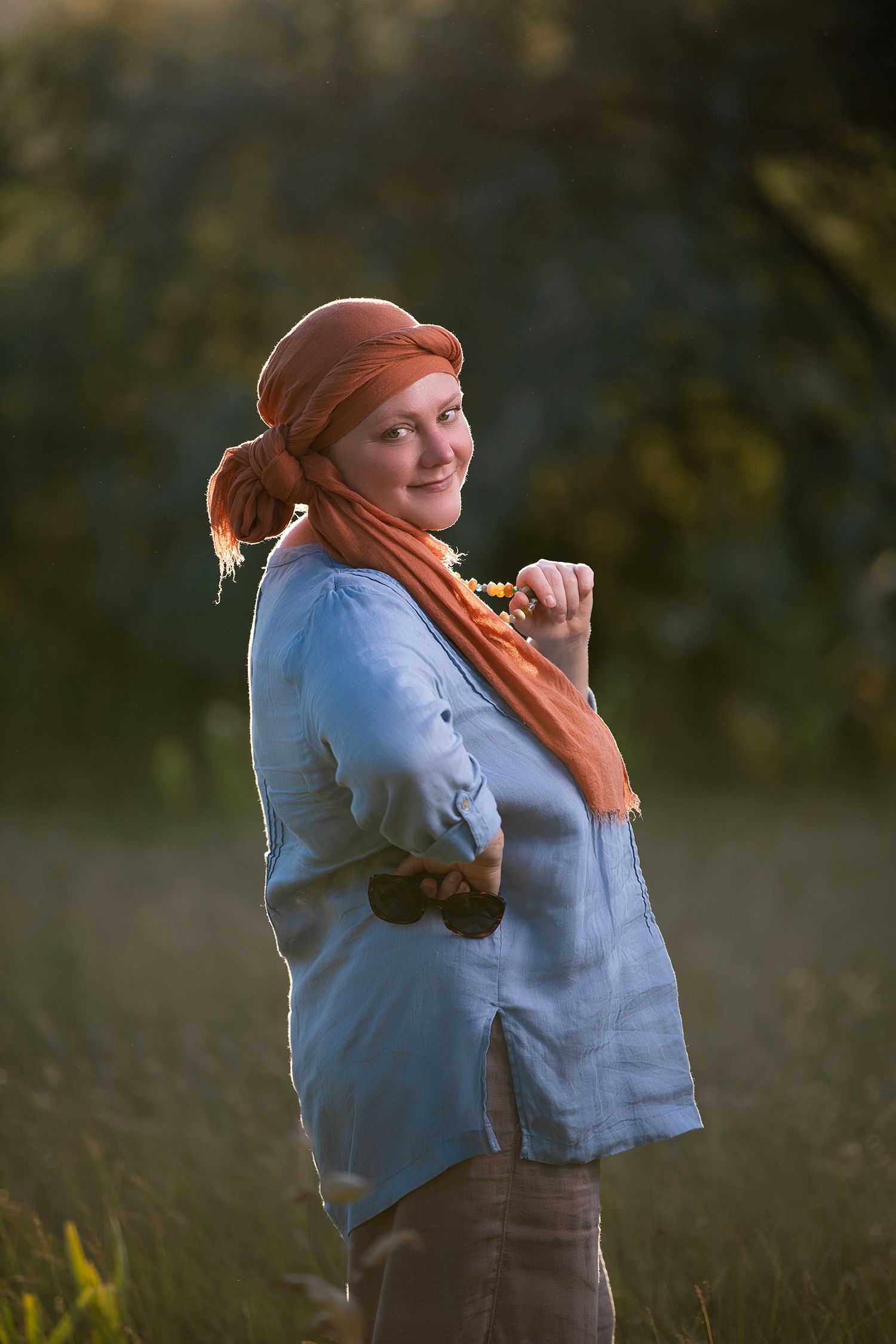 Woman in rust head wrap and light blue shirt smiling in side profile, holding necklace and sunglasses - Okanagan portrait by Tania Veitch.