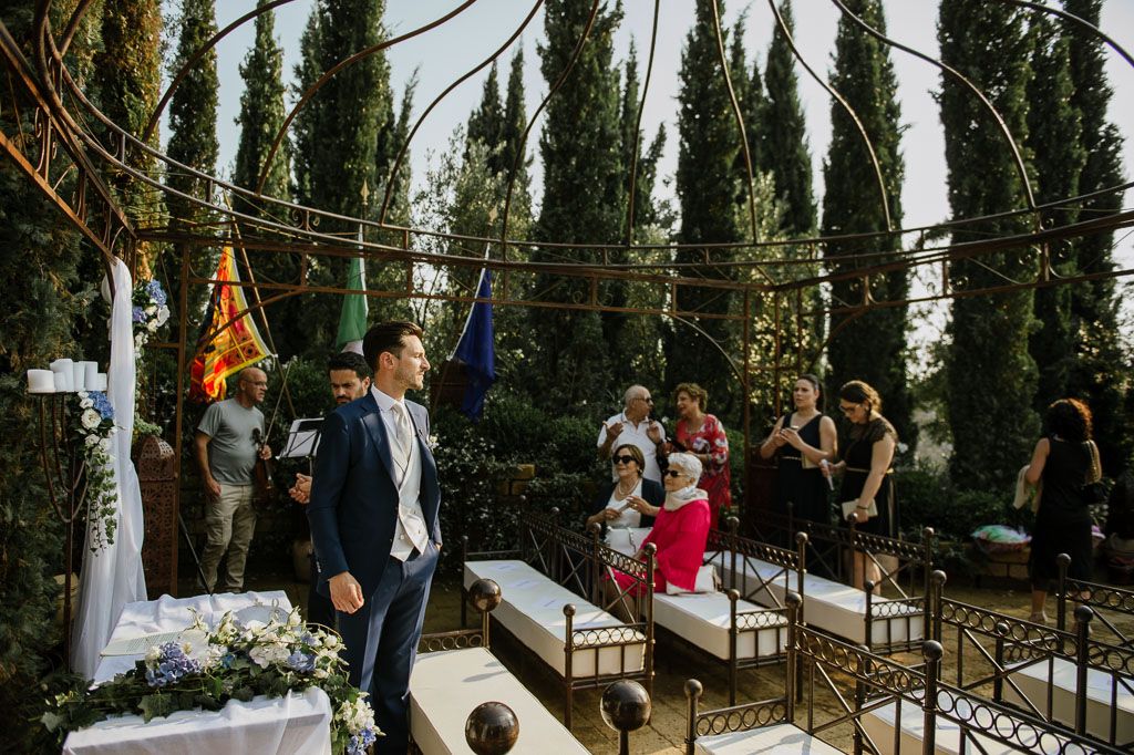 Outdoor wedding ceremony setup with wooden chairs and floral arrangements between cypress trees.