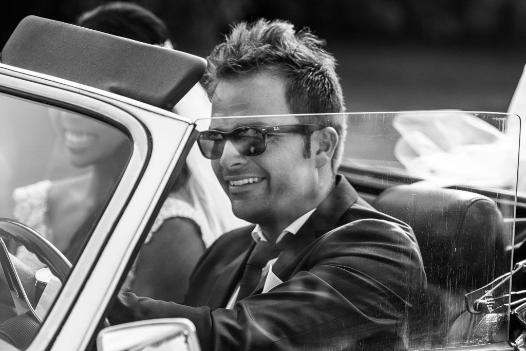 A black and white candid photo shows the bridal party sharing laughter in a vintage car on a wedding day.