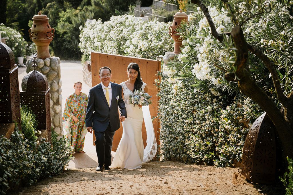 A romantic wedding couple walks through a garden archway with white flowering vines and terracotta details.