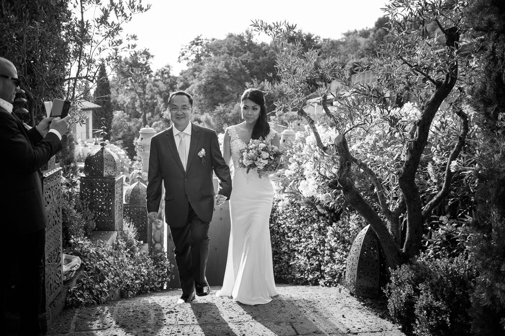 Black and white photo of newlyweds walking down a scenic garden path surrounded by trees.