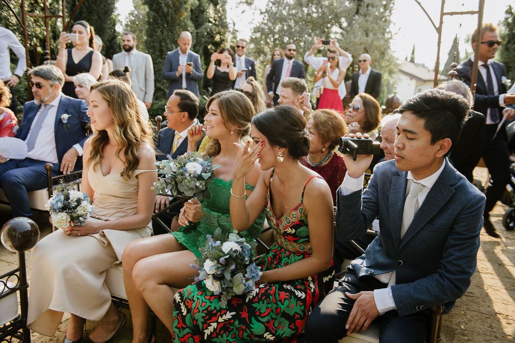 Wedding ceremony guests seated outdoors in summer attire with decorative floral arrangements.