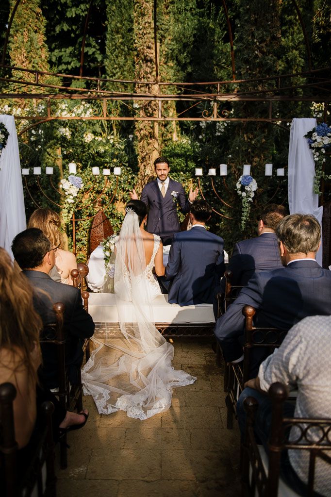 Outdoor wedding ceremony setup with white decor and guests seated in a wooded setting.