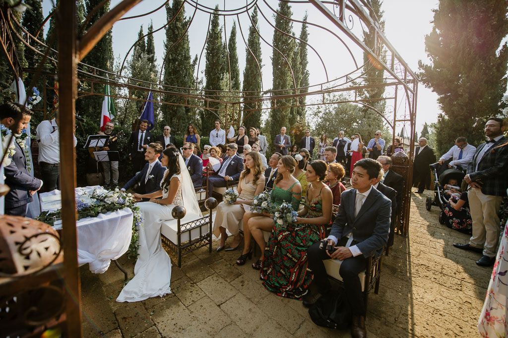 Outdoor wedding ceremony with guests seated beneath cypress trees and decorative lighting.
