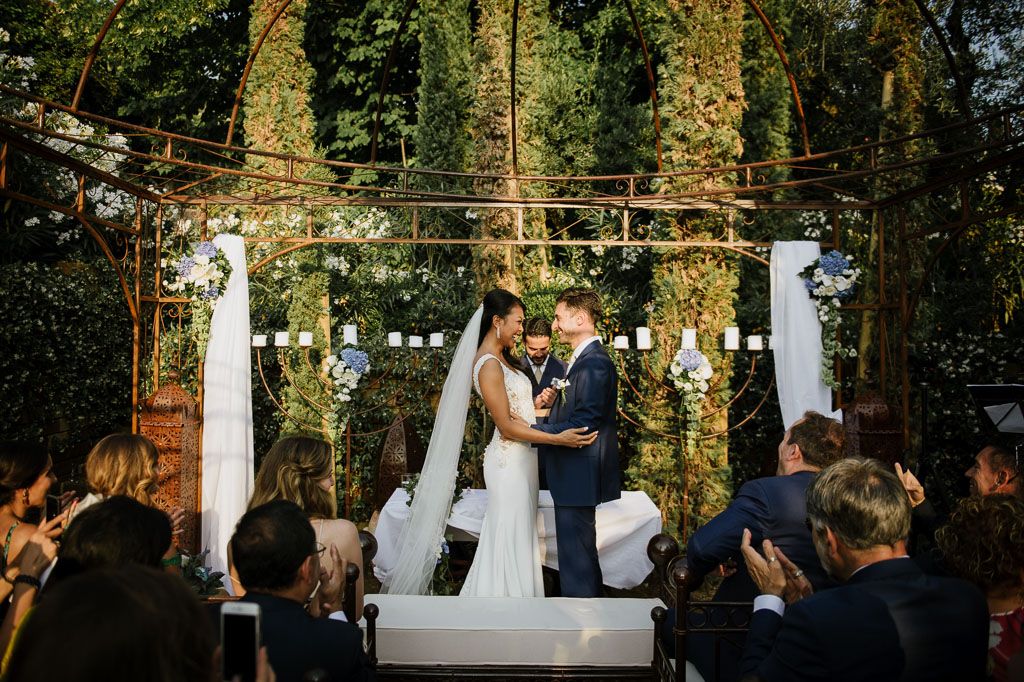 Outdoor forest wedding ceremony with couple at decorated altar under string lights and greenery.