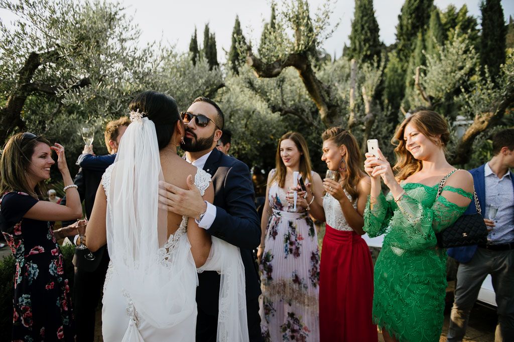 Wedding guests mingling outdoors among olive trees during a daytime celebration.