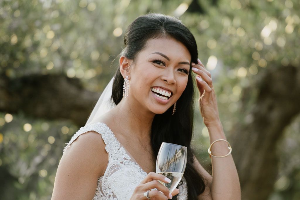 Bride in white dress holding champagne glass and smiling in natural outdoor lighting.