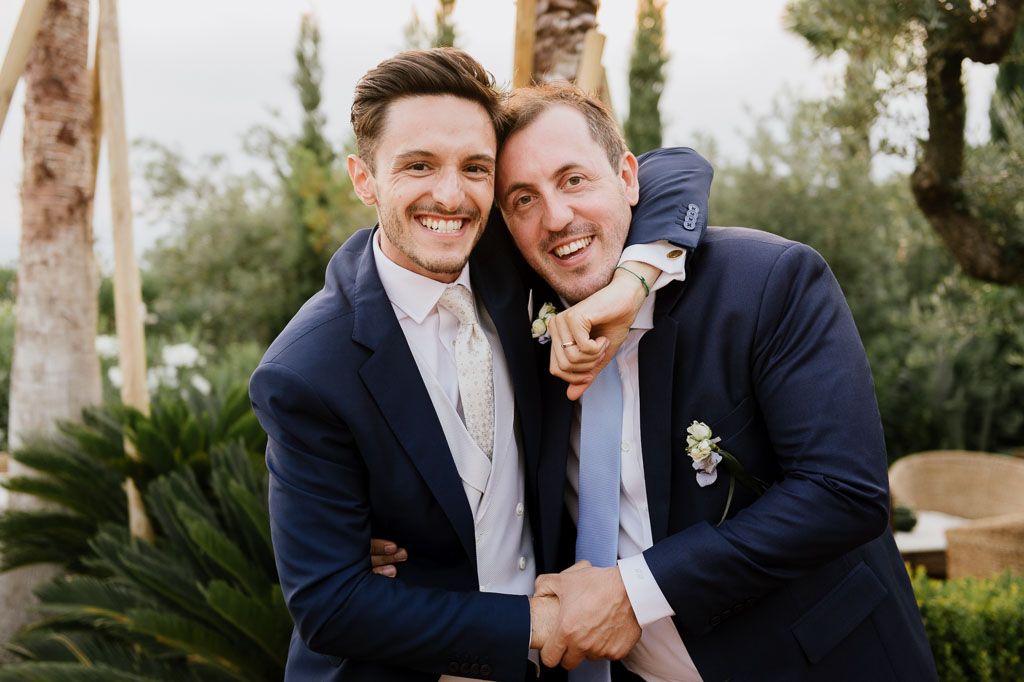 Two grooms in formal wedding attire sharing a joyful moment together outdoors.