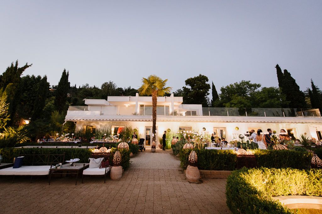 Elegant white villa venue illuminated at dusk with palm trees and outdoor reception setup.