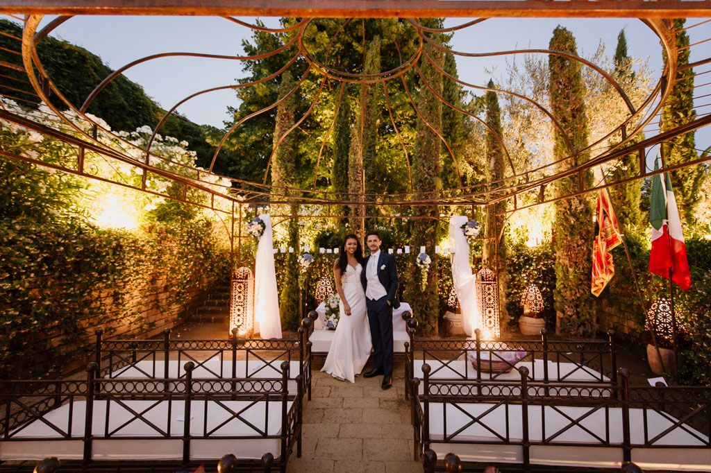 Wedding ceremony in a stunning garden setting with string lights and cypress trees at sunset.