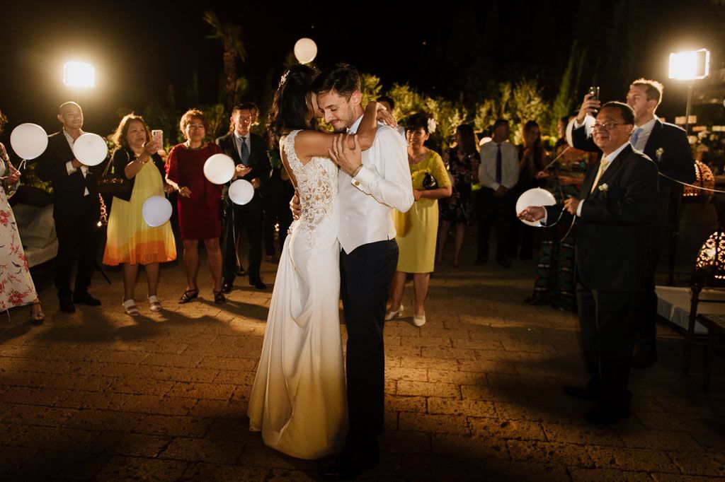 Wedding guests celebrate on a dance floor at night illuminated by string lights and paper lanterns.