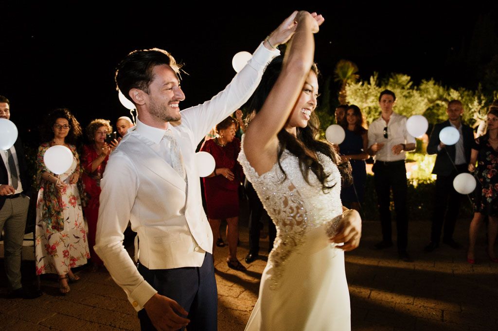 Couple dances together during an outdoor evening reception with twinkling lights and guests in the background.