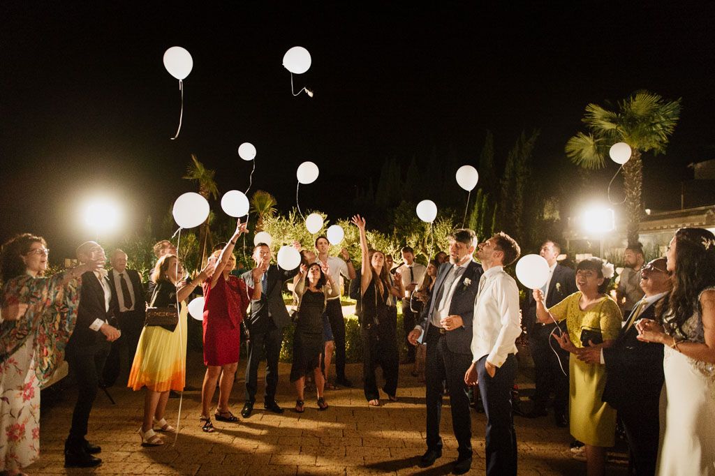White balloons float above dancing party guests during a nighttime celebration.