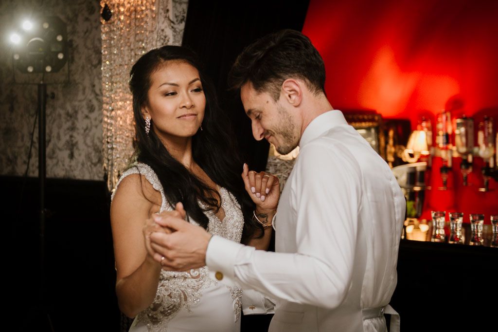 Wedding couple share a romantic dance in a dimly lit reception venue with red ambient lighting and crystal chandeliers.