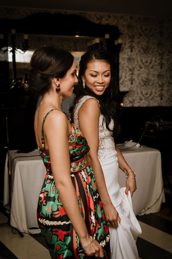 Two friends share a joyful dance moment at a wedding reception, one wearing a floral dress and one in white.