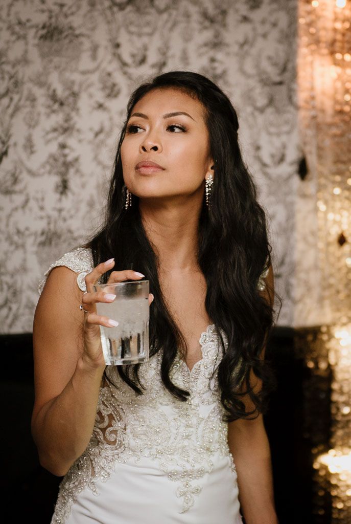 A bride in an elegant lace dress holds a drink while posing against a textured wall at her reception.