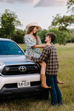 A romantic couple leans against a Toyota RAV4 with Texas plates in a grassy field during golden hour.