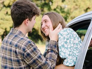A couple shares a romantic moment as they lean against a white car door in casual outdoor attire.