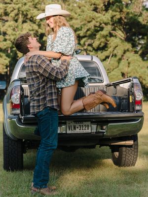 Romantic couple sits together on a black pickup truck tailgate in a sunny outdoor setting.