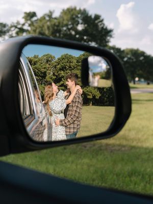 A romantic moment reflected in a car's side mirror, showing embracing figures against a green grassy background.