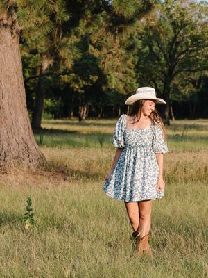 A floral dress-wearing figure stands in a sunny meadow with pine trees in the background.