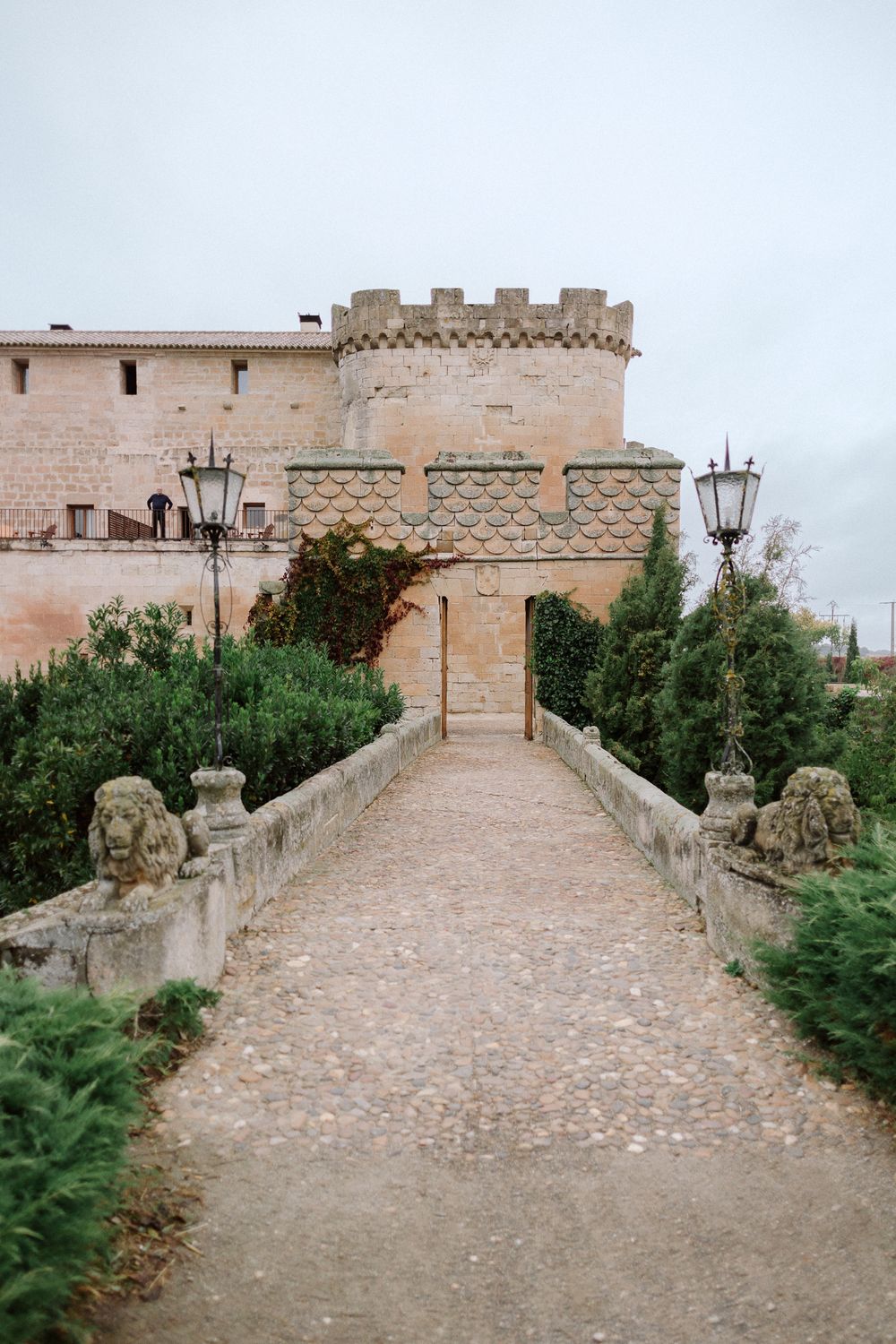 Stone bridge pathway leads to a medieval castle entrance with crenellated tower and decorative lampposts in Spain.