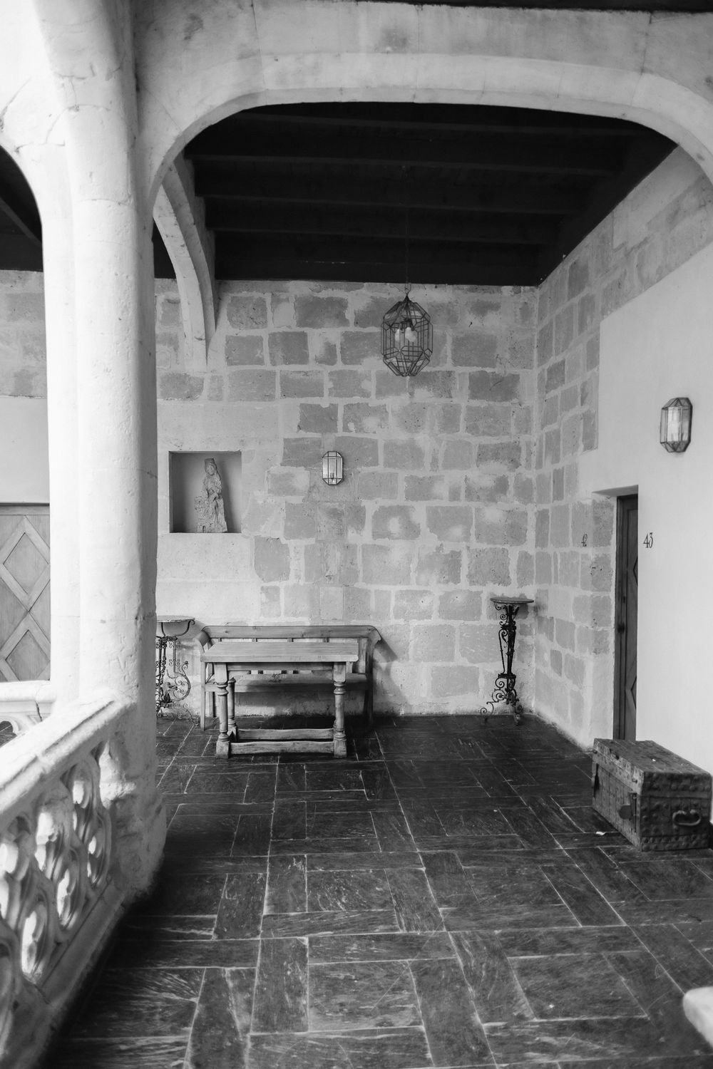 A stone-walled medieval corridor with hanging lanterns, slate flooring, and arched ceiling features a small altar space.