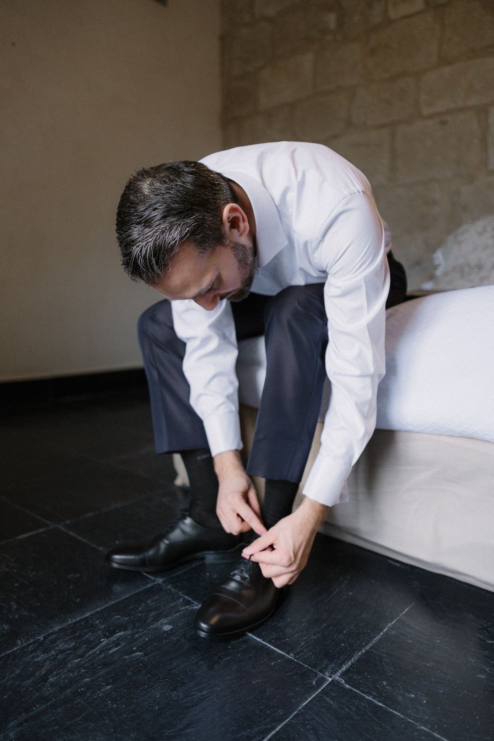 Someone in formal attire ties their black leather dress shoes while sitting on a white sofa against a stone wall.