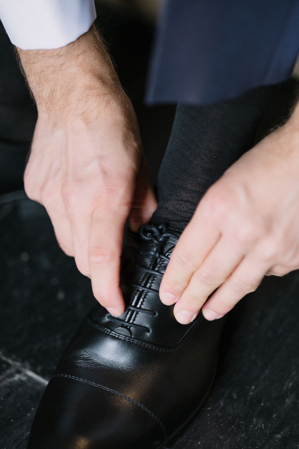 Close-up of hands tying laces on a black leather dress shoe against dark background.