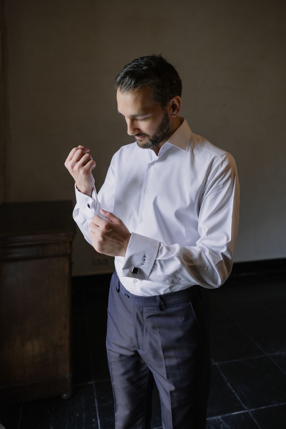 Person in white dress shirt and dark trousers adjusts their sleeve cuff in a moody indoor setting.
