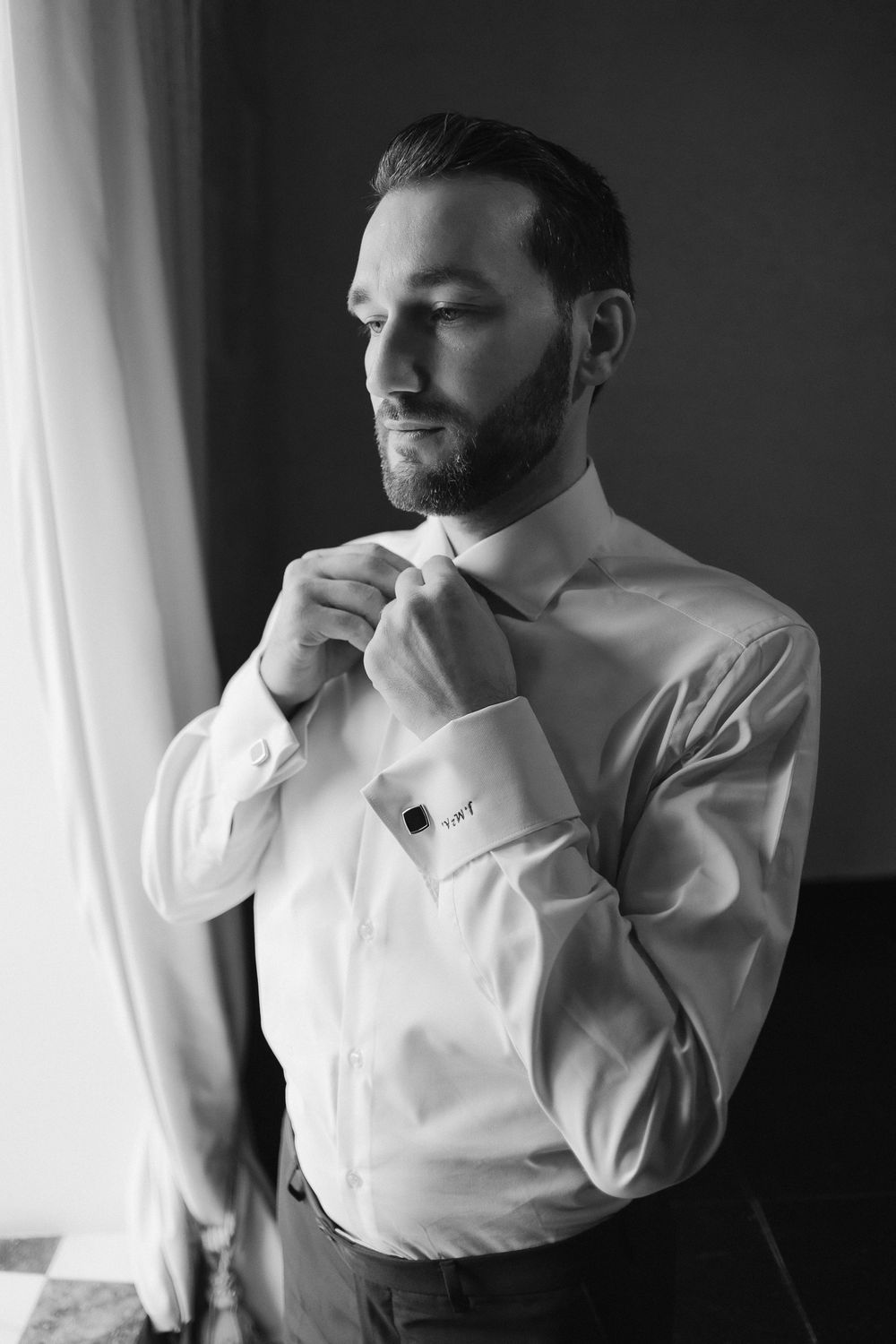 Black and white photo of groom adjusting his tie by a window in formal white dress shirt and black pants.