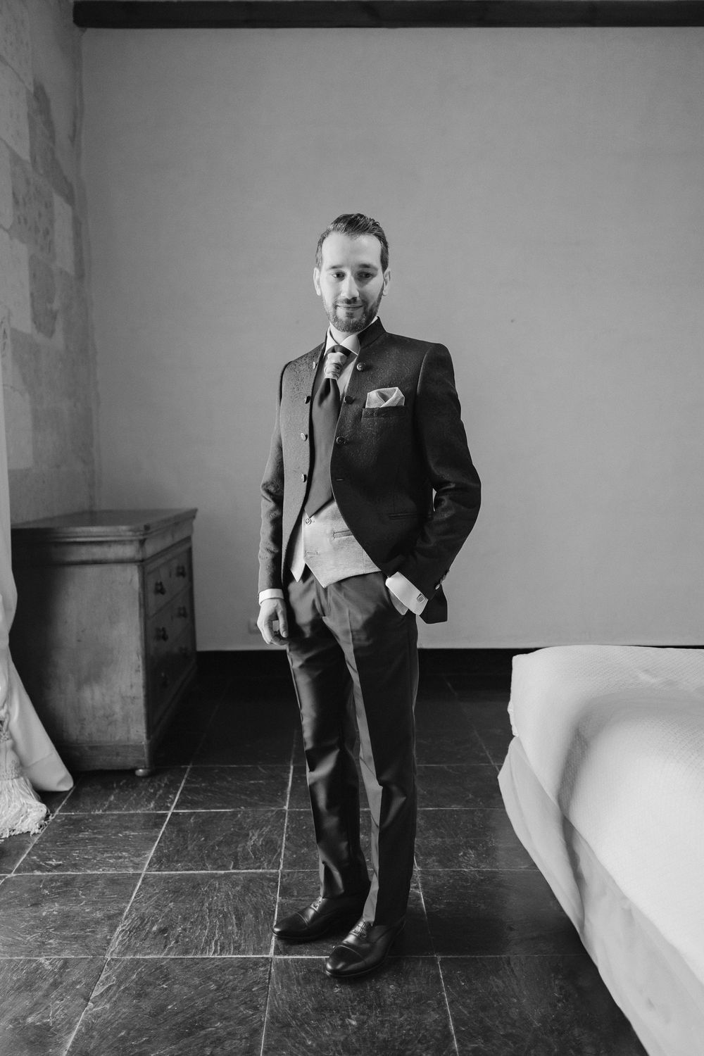 A black and white photograph of someone in a formal suit standing in an interior space with stone flooring.