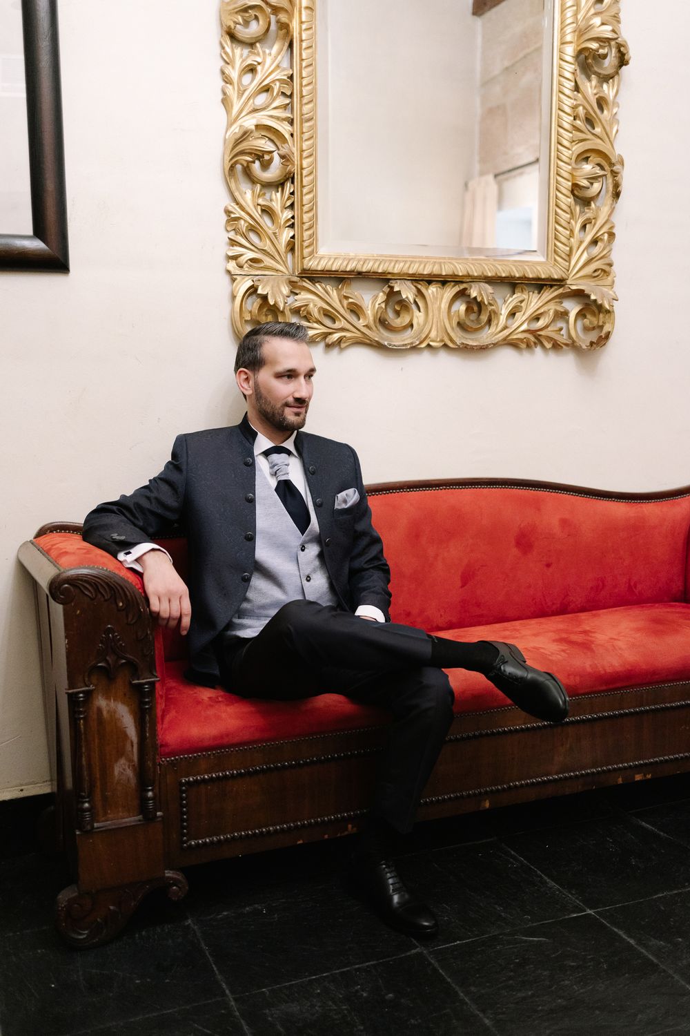 A groom in a black tuxedo reclines on a vintage red velvet sofa beneath an ornate gold-framed mirror.