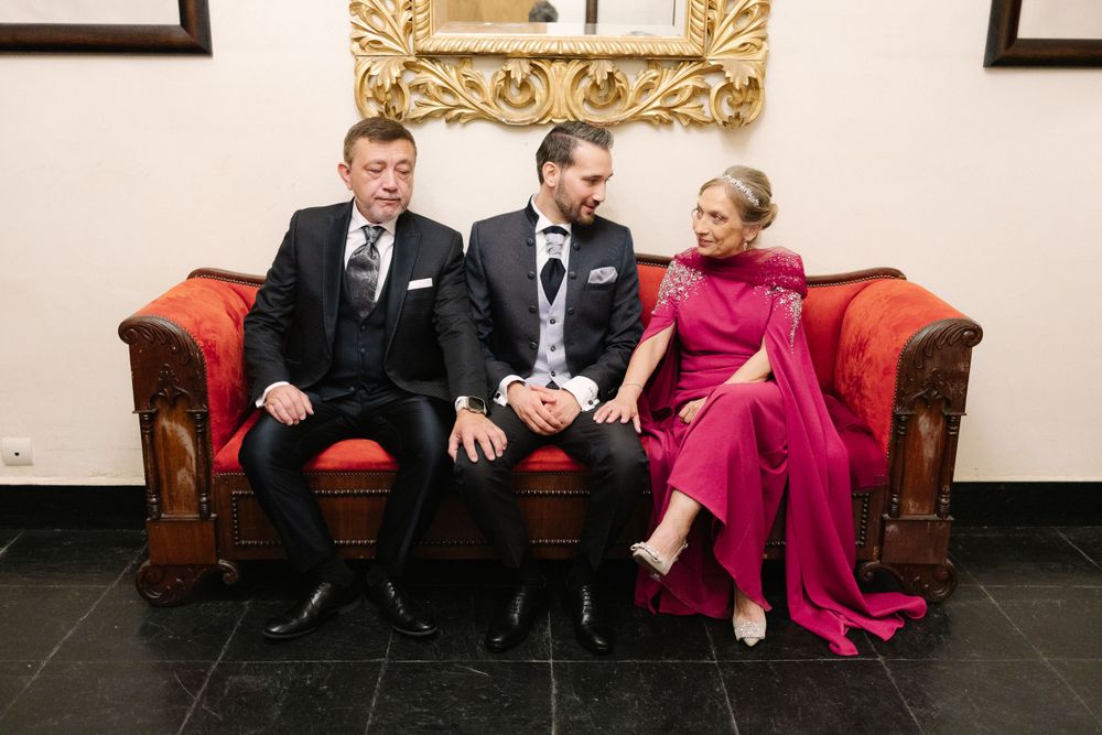 Three people in formal attire sit together on a red leather couch, with one wearing a vibrant pink dress.