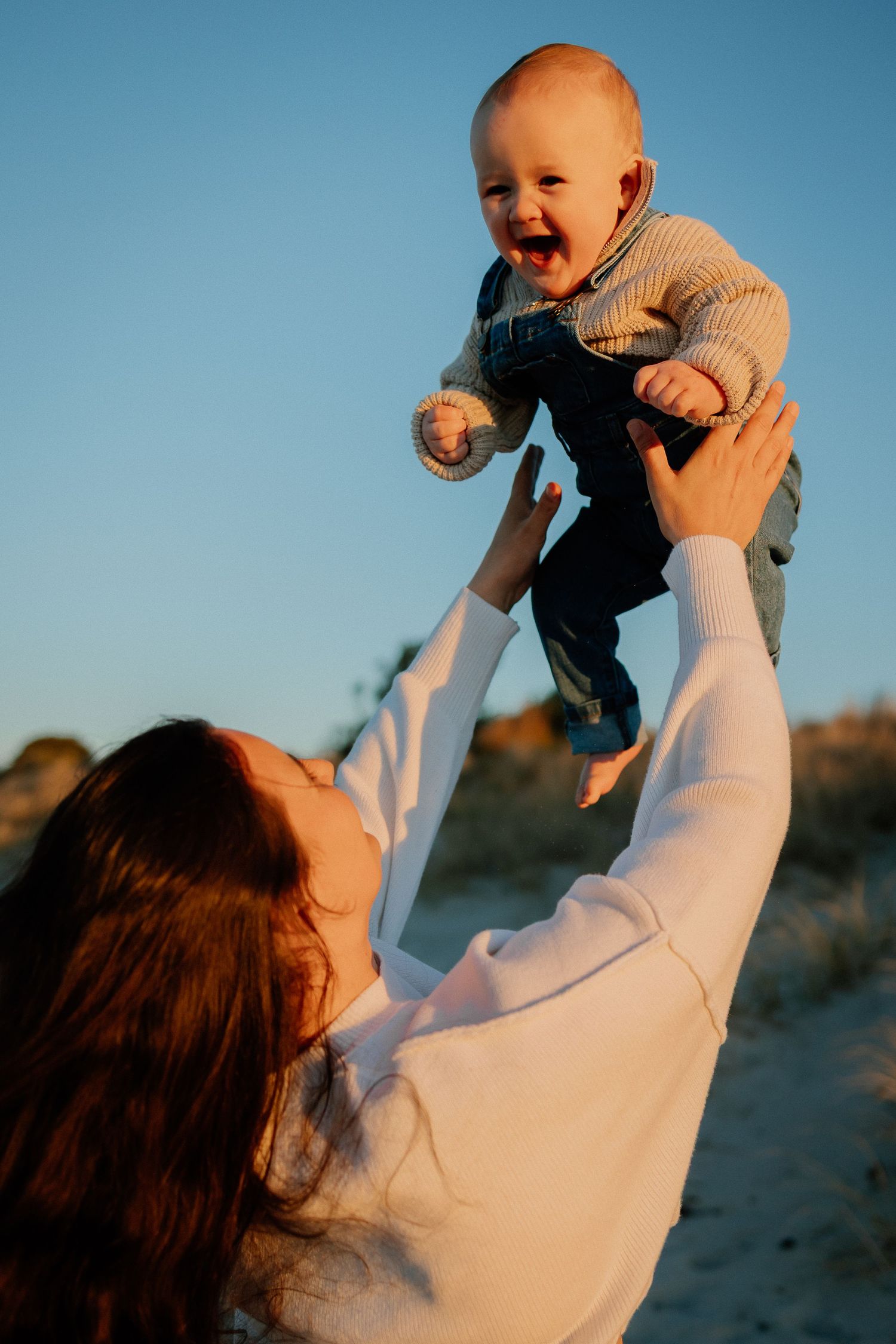 A baby wearing a sweater and jeans being tossed in the air against a blue sky, expressing pure joy and laughter.