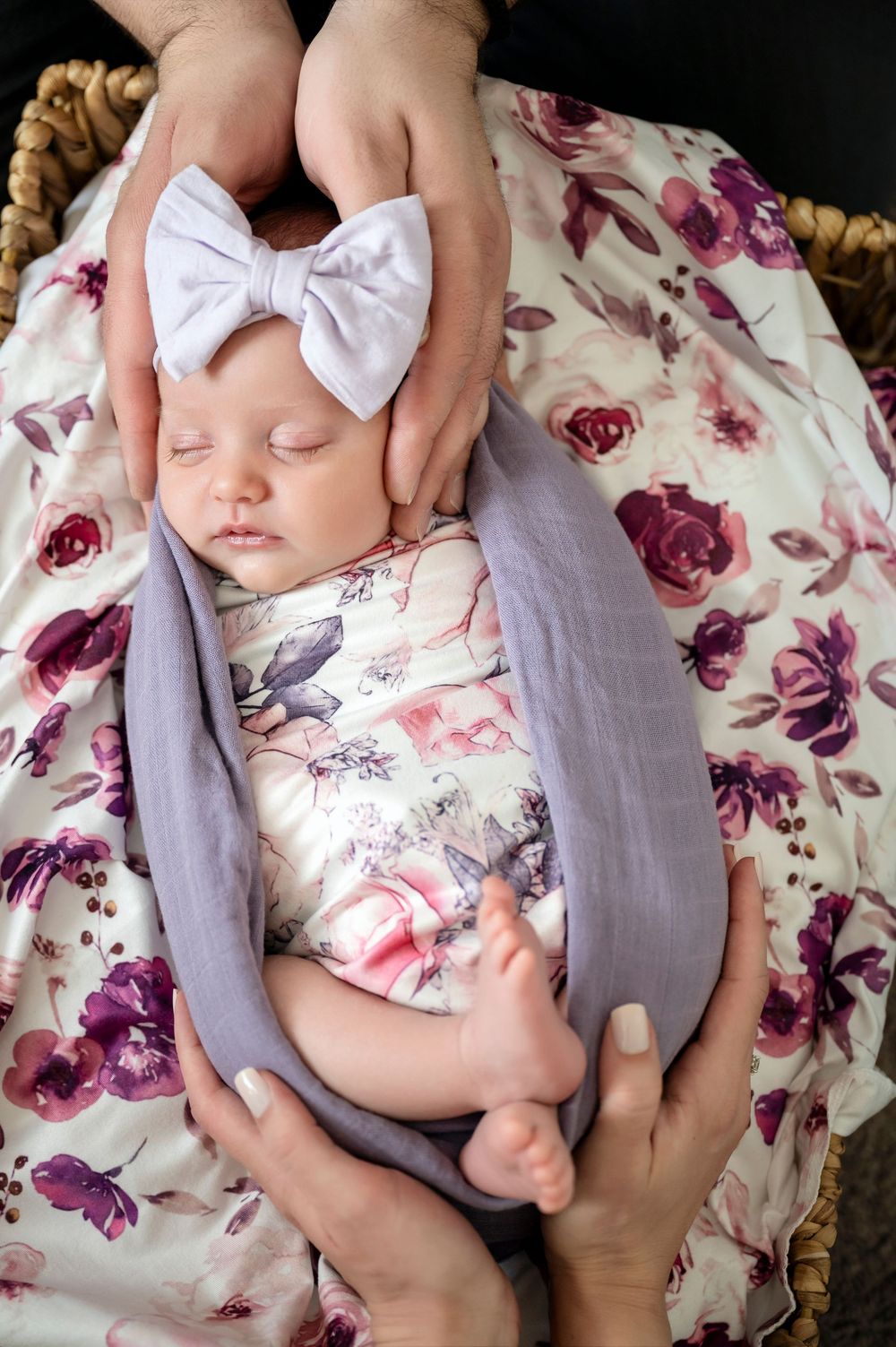 Sleeping baby wearing white bow headband lies on floral bedding with purple roses while hands gently touch.