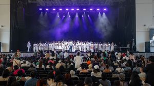 Large choir performing on stage with purple lighting and audience seated in foreground.