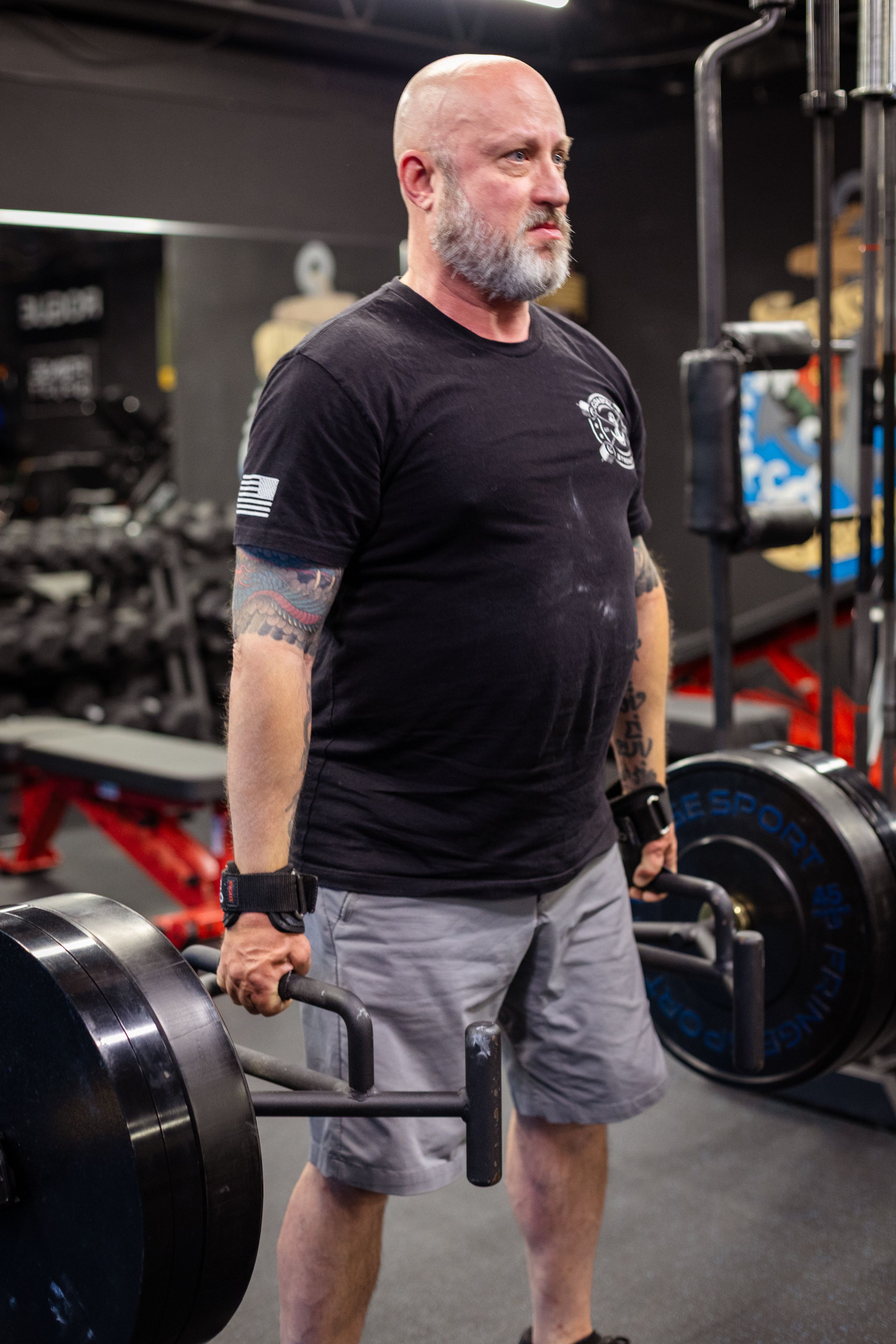 Bearded man in black t-shirt and shorts standing with weights at gym.