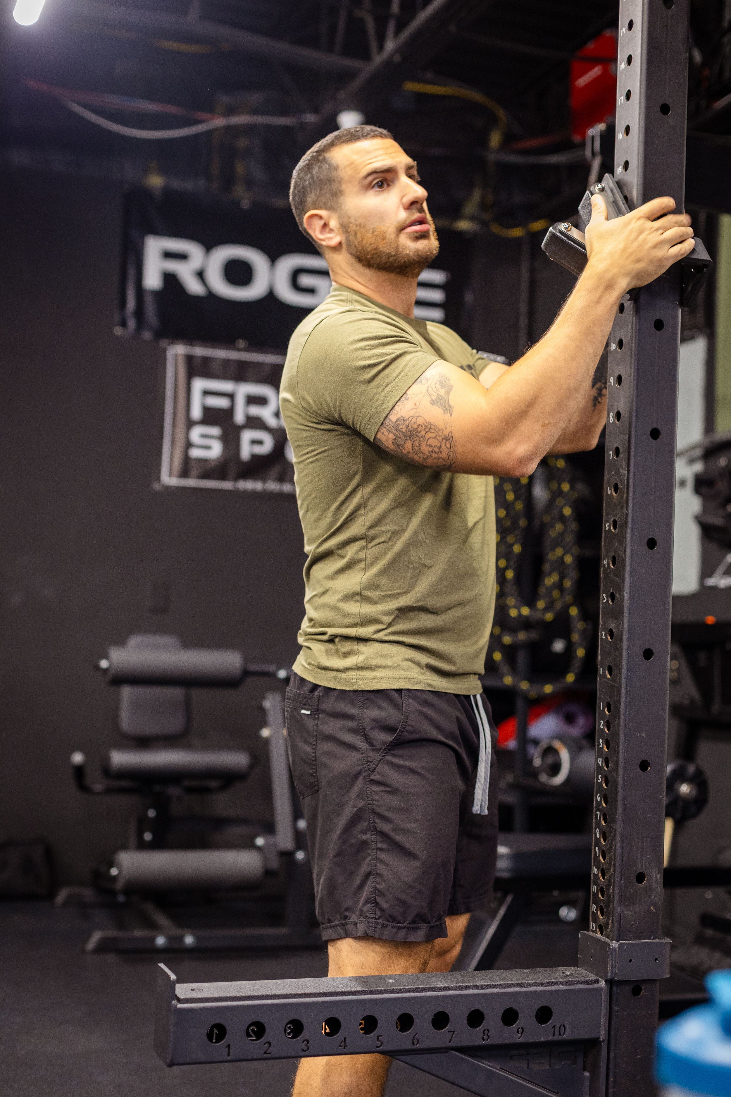 Athletic man in olive shirt adjusting equipment on power rack at fitness center.