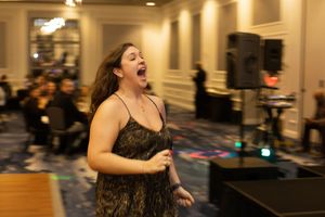 A performer in a black lace dress sings passionately during an indoor event.