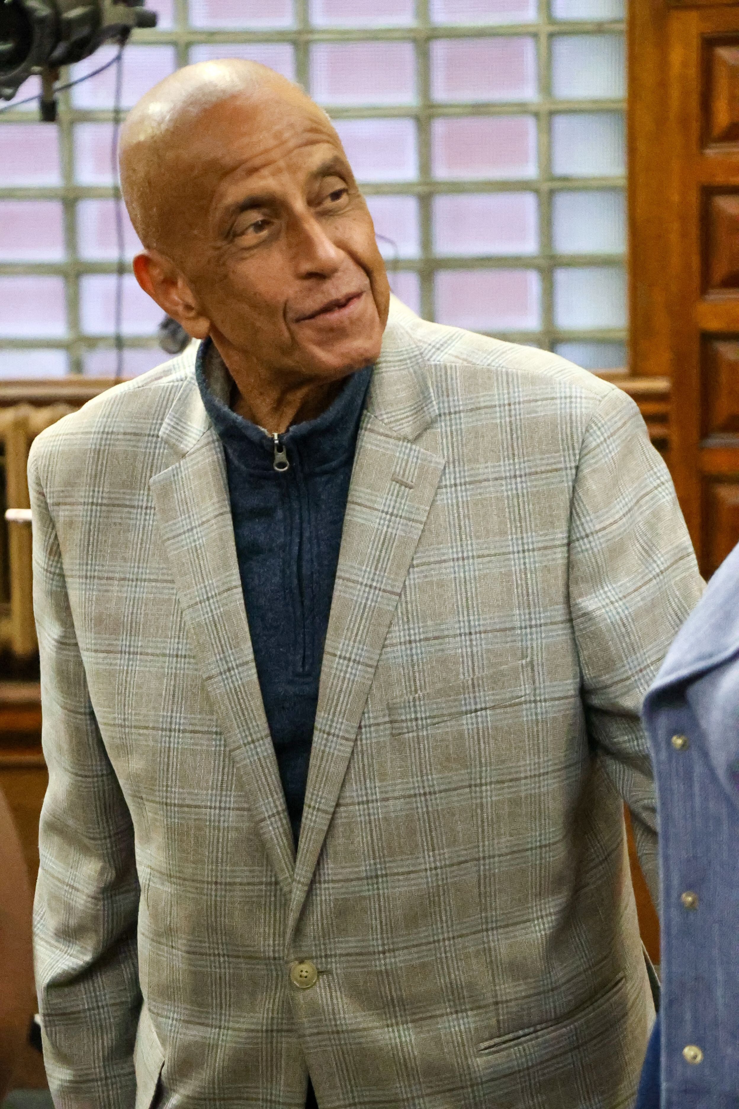 Elderly bald man in plaid blazer and navy sweater smiling indoors.